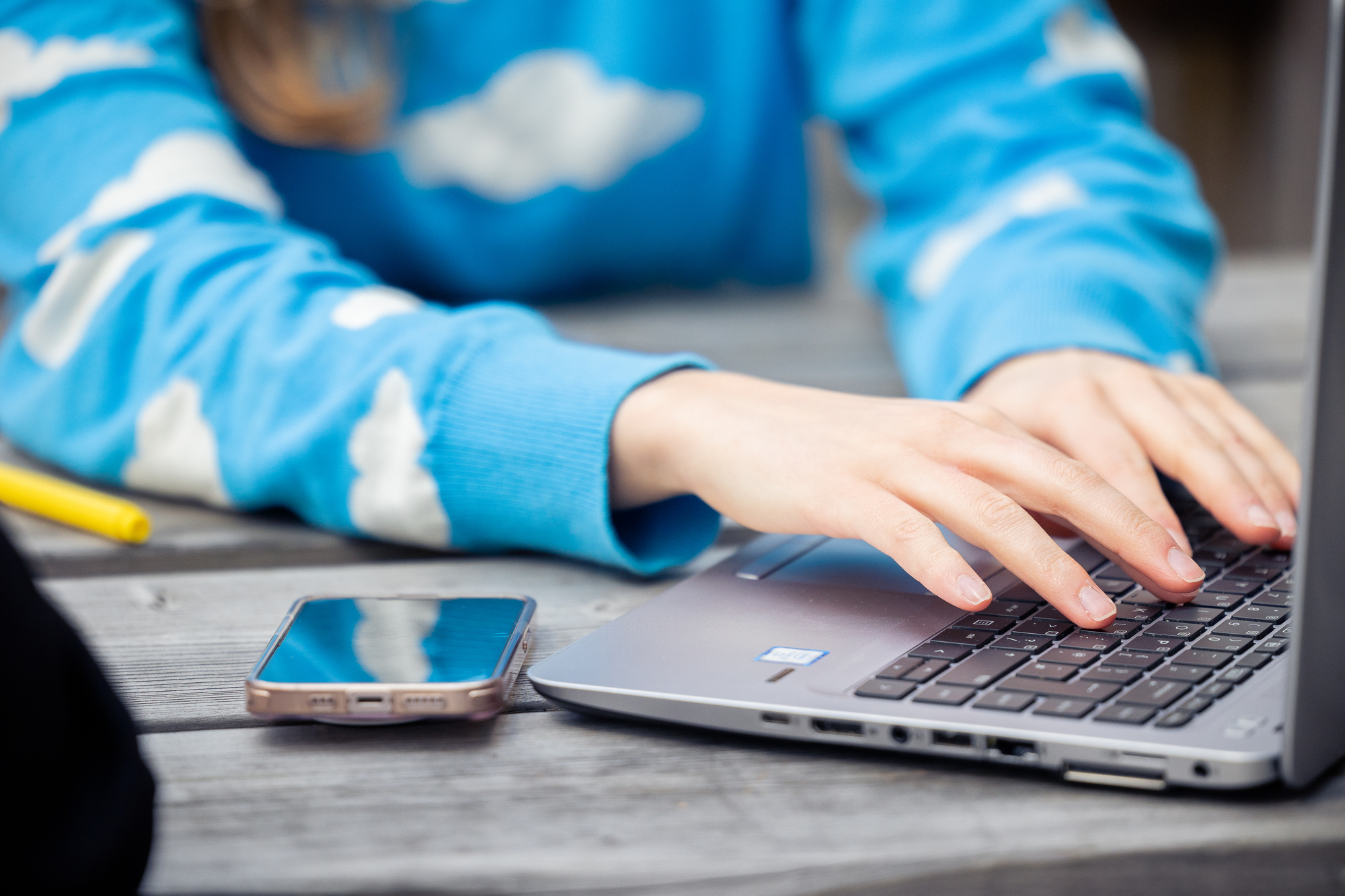 close up of two white hands typing onto a laptop on a bench. 