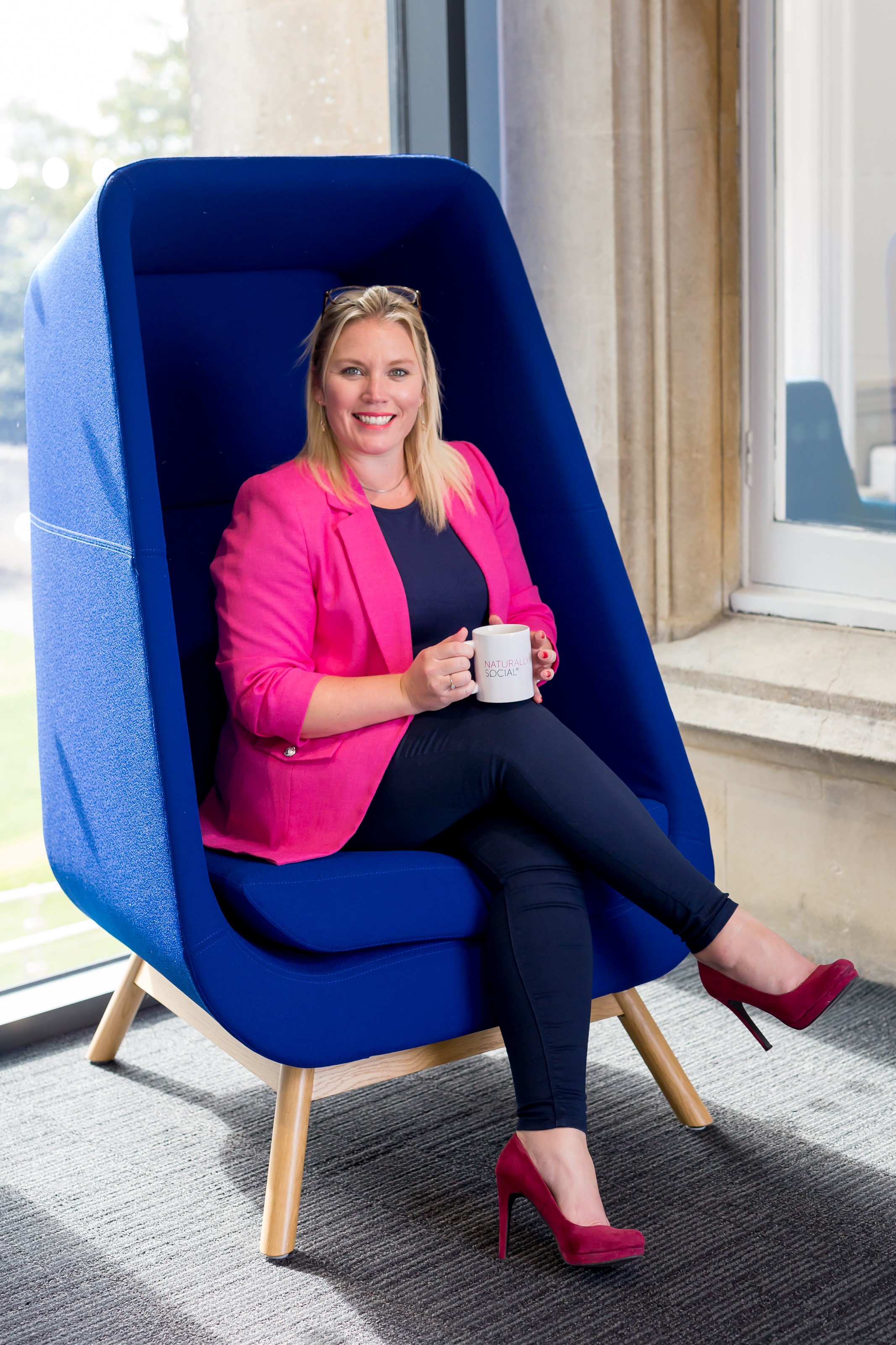 Woman with blond hair sat in a Blue chair wearing a pink jacket, blue top, blue jeans, pink high heals. She is smiling and holding a mug that says Naturally Social on it
